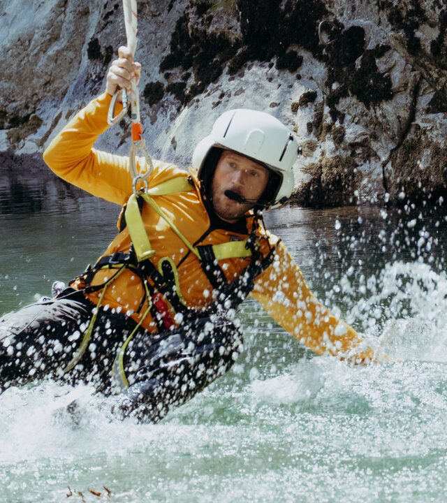 "Die Bergretter: Ohne Ausweg": In einem Wildbach: Bergretter Markus Kofler (Sebastian Ströbel) hängt am Rettungstau. Seine Beine sowie der linke Arm sind ins aufwirbelnde Wasser getaucht. Er blickt konzentriert.