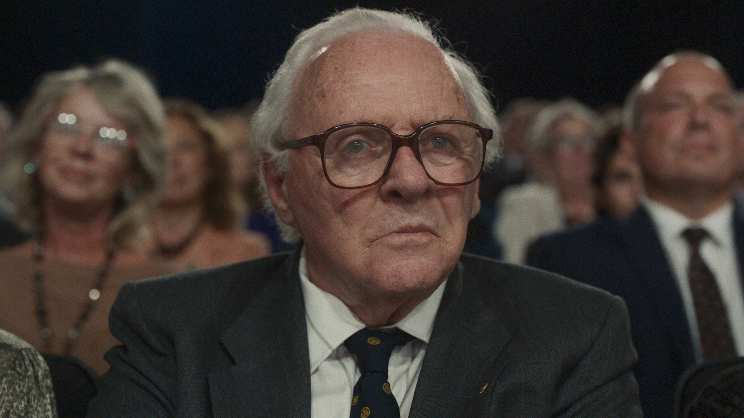 "One Life - Wer ein Leben rettet, rettet die ganze Welt": Nahaufnahme: Nicholas Winton (Anthony Hopkins) mit großer Brille, weißem Haar, in Anzug und Krawatte, sitzt im Publikum einer TV-Show in der ersten Reihe und blickt etwas skeptisch nach vorne.