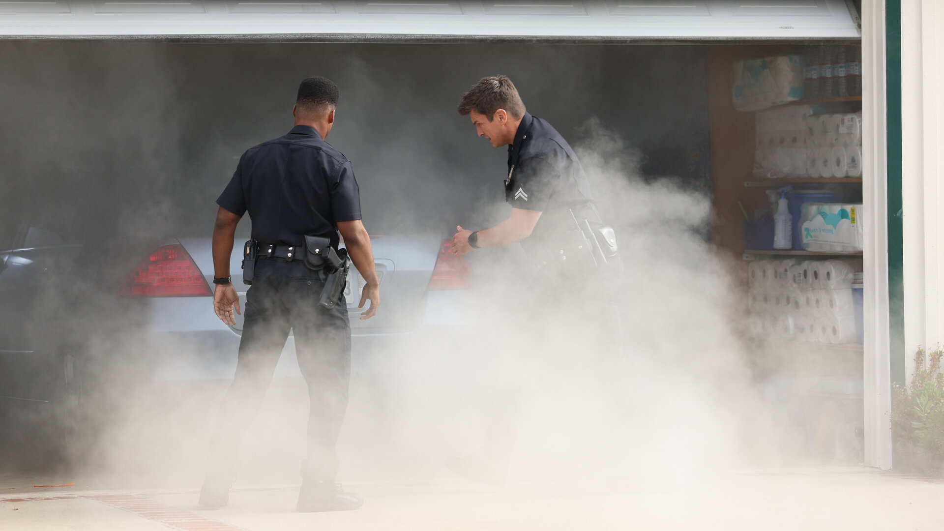 "The Rookie - Neue Wege": Officer Nolan (Nathan Fillion) und Officer Aaron Thorsen (Tru Valentino) stehen vor einer Garage, aus der dichte Schwaden von Rauch hervorquellen.