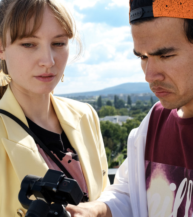 "SOKO Wien - Nahaufnahme": Auf einem Balkon stehen Dani (Julia Roy) und Flo (Bagher Ahmadi). In einer Hand hält Flo Neubauer eine Kamera, auf die beide Protagonisten schauen.