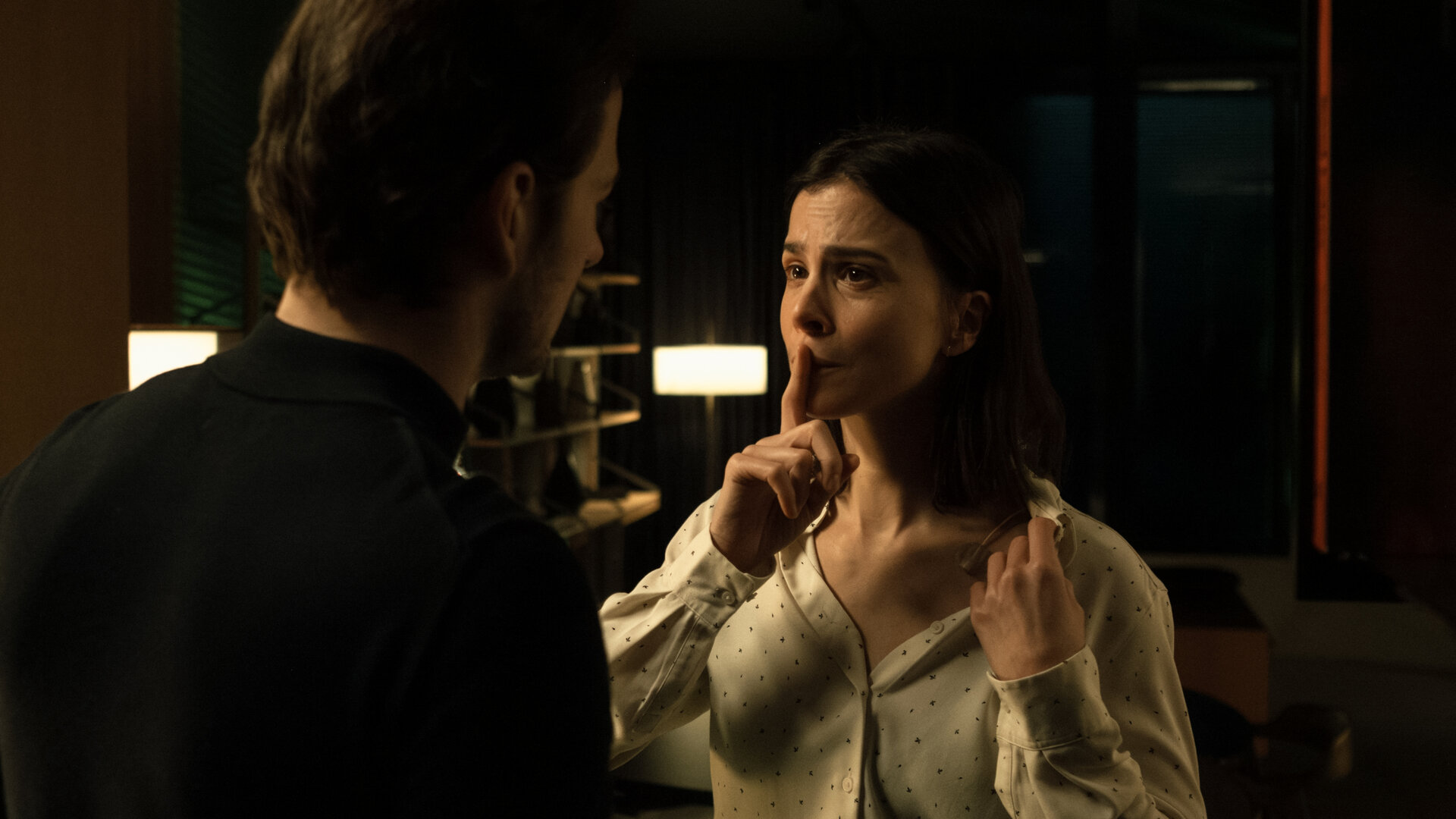 "Serial Cleaner - Kollisionskurs": In einem dunklen Innenraum stehen sich Teri Marquard (Aylin Tezel) und Vincent Karanz (Stefan Gorski) gegenüber, Tischleuchten im Hintergrund spenden spärliches Licht. Teri hält ihren Zeigefinger an ihren Mund, damit Vincent keinen Ton von sich gibt. Sie zeigt Vincent, dass sie auf der linken Brustseite verkabelt ist.