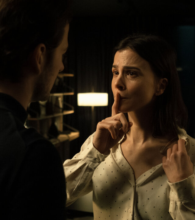"Serial Cleaner - Kollisionskurs": In einem dunklen Innenraum stehen sich Teri Marquard (Aylin Tezel) und Vincent Karanz (Stefan Gorski) gegenüber, Tischleuchten im Hintergrund spenden spärliches Licht. Teri hält ihren Zeigefinger an ihren Mund, damit Vincent keinen Ton von sich gibt. Sie zeigt Vincent, dass sie auf der linken Brustseite verkabelt ist.