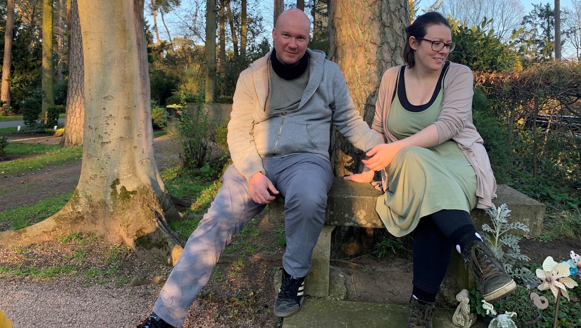 "37° Ein Hauch von Leben - Sternenkinder und ihre Eltern": Auf einem Friedhofsgelände sitzen Lydia und Dietmar nebeneinander unter einem großen Baum auf einer Steinbank .