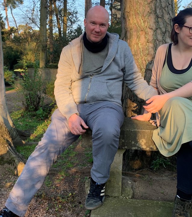 "37° Ein Hauch von Leben - Sternenkinder und ihre Eltern": Auf einem Friedhofsgelände sitzen Lydia und Dietmar nebeneinander unter einem großen Baum auf einer Steinbank .