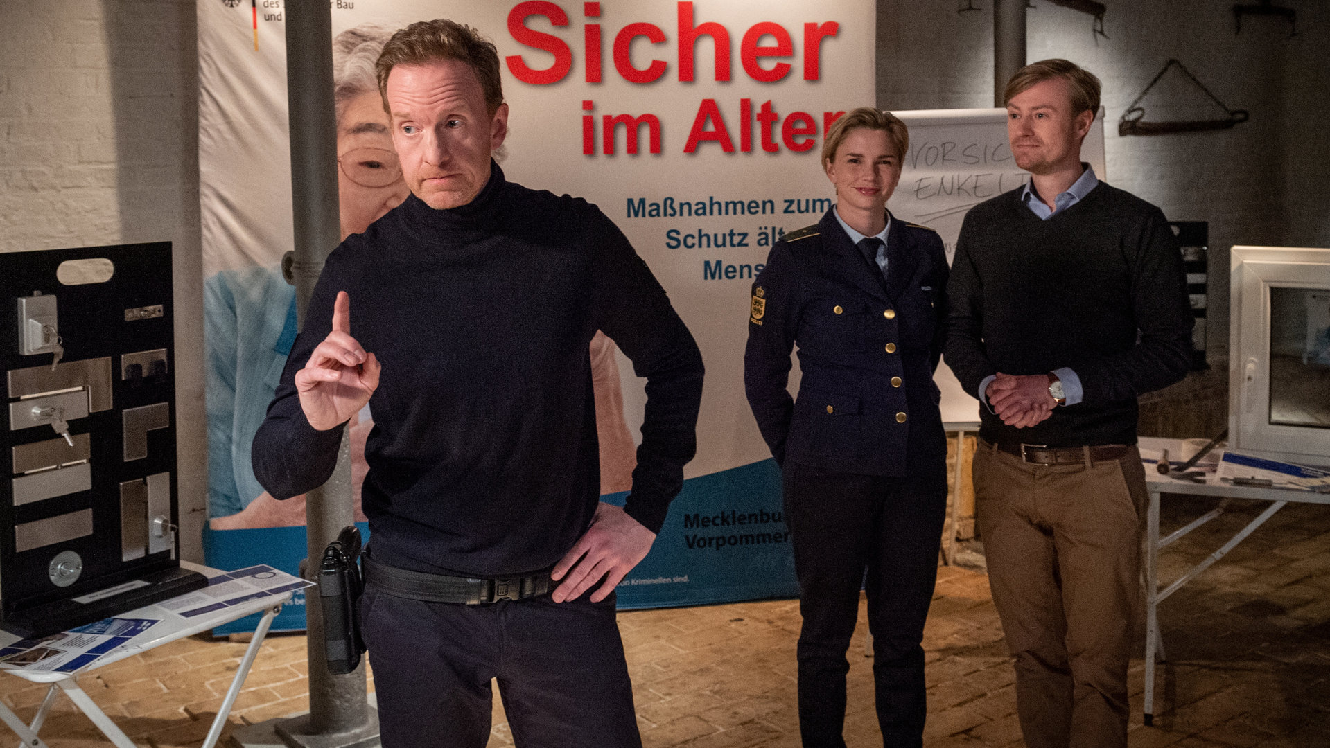 „SOKO Wismar – Sicher im Alter“: Timmermann (Mathias Junge) steht mit erhobenem Zeigefinger inmitten eines großen Saales. Hinter ihm stehen Stine (Sidsel Hindhede) und Felix Brechtel (Eric Bouwer). Im Hintergrund hängt ein großes Banner mit der Aufschrift "Sicher im Alter – Maßnahmen zum Schutz älterer Menschen". Auf einem Beistelltisch sind verschiedene Schlösser ausgestellt. Auf einem weiteren Tisch steht ein Fenster zu Demonstrationszwecken.