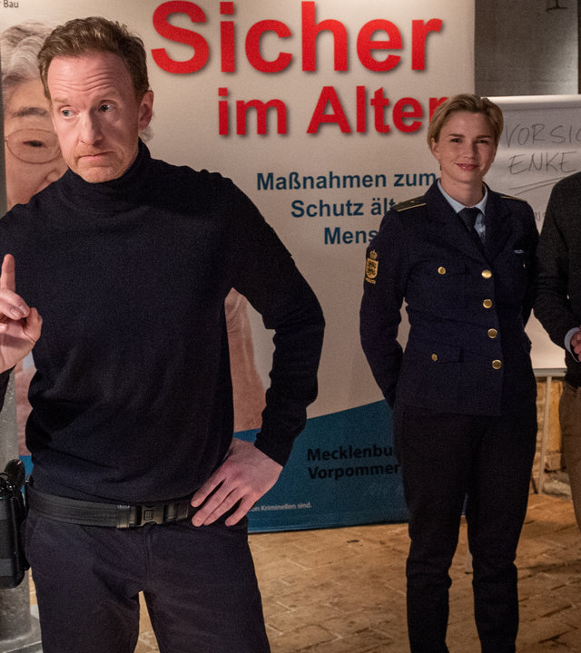 „SOKO Wismar – Sicher im Alter“: Timmermann (Mathias Junge) steht mit erhobenem Zeigefinger inmitten eines großen Saales. Hinter ihm stehen Stine (Sidsel Hindhede) und Felix Brechtel (Eric Bouwer). Im Hintergrund hängt ein großes Banner mit der Aufschrift "Sicher im Alter – Maßnahmen zum Schutz älterer Menschen". Auf einem Beistelltisch sind verschiedene Schlösser ausgestellt. Auf einem weiteren Tisch steht ein Fenster zu Demonstrationszwecken.
