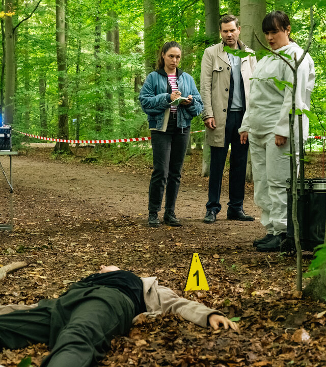 "SOKO Köln - Mords-Polka": Nadja Schilling (Kaja Schmidt-Tychsen) liegt mit ausgestreckten Armen und Beinen in Rückenlage in einem Wald auf dem Boden. Hinter ihr stehen neben einem Baum Amira Mahdi (Aylin Ravanyar), Jonas Fischer (Lukas Piloty) und Dr. Westphal (Yulia Yáñez Schmidt) nebeneinander. Amira sieht ernst zu Dr. Westphal. Die Rechtsmedizinerin trägt einen weißen Schutzanzug und blickt ernst zur Toten. Jonas schaut ebenfalls zu Nadja Schilling.