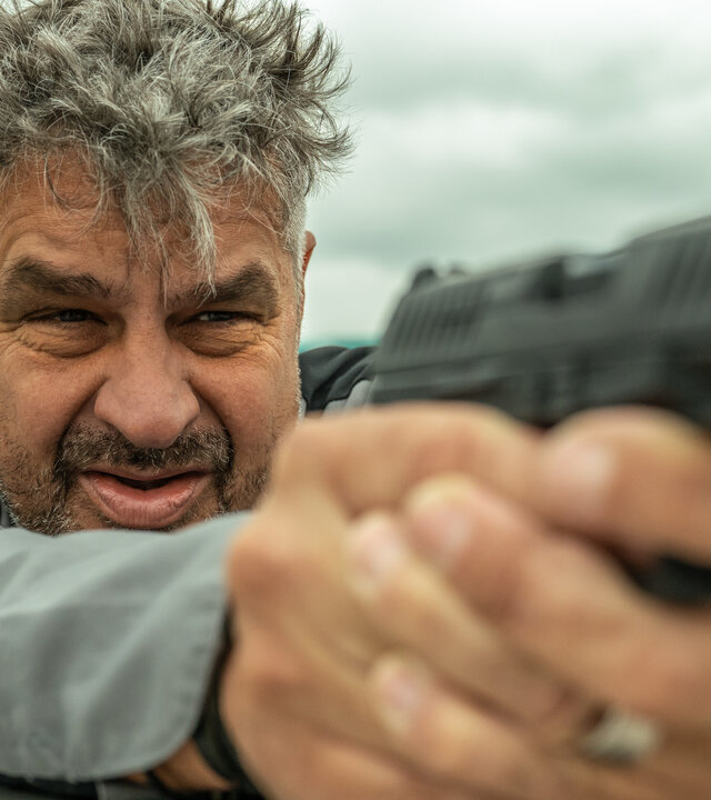 "Blind Sherlock - Pfannkuchenschiff": Nico (Frank Lammers) steht auf einem Boot. Er hält seine Pistole mit beiden Händen und visiert ein Ziel an.