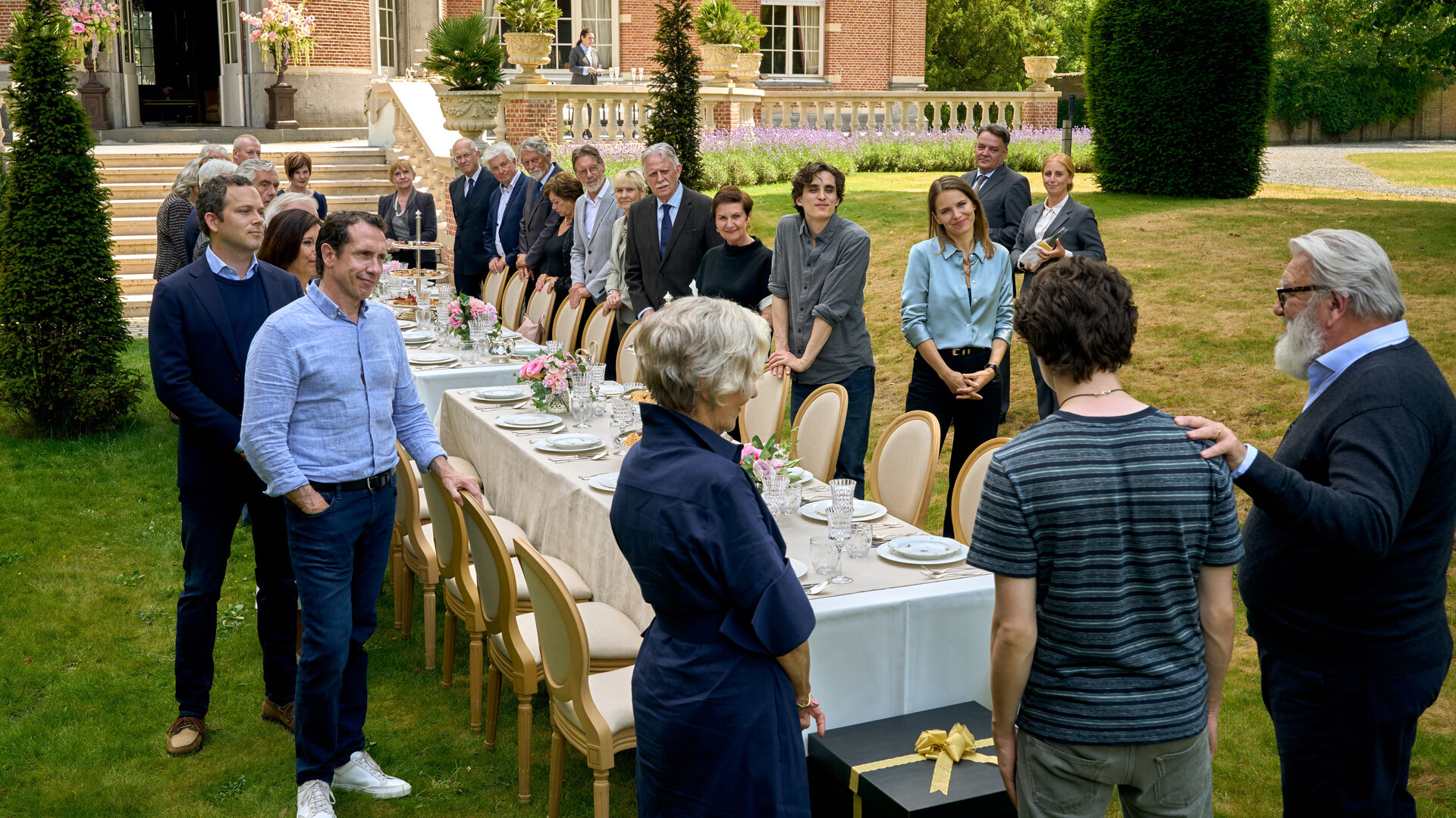 "Elixir – The Pain Killers - Die Auserwählten": In einem großen Garten vor einem schlossartig aussehenden Gebäude sind zwei lange, festlich gedeckte Tische aufgestellt. Im Vordergrund sind Riekje (Katelijne Damen), Art (Roman Derwig) und Ludo (Wim Opbrouck) von hinten zu sehen. Ihnen gegenüber stehen zahlreiche Gäste an den Tischen. Vor Art steht ein großes Geschenkpaket auf dem Boden.