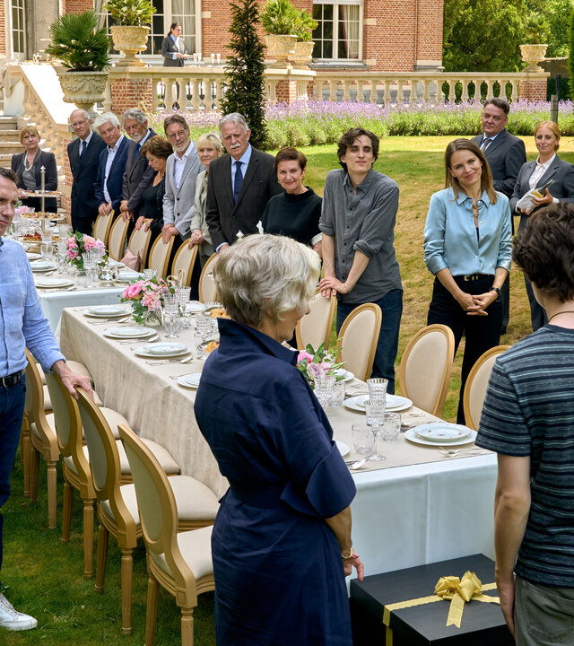 "Elixir – The Pain Killers - Die Auserwählten": In einem großen Garten vor einem schlossartig aussehenden Gebäude sind zwei lange, festlich gedeckte Tische aufgestellt. Im Vordergrund sind Riekje (Katelijne Damen), Art (Roman Derwig) und Ludo (Wim Opbrouck) von hinten zu sehen. Ihnen gegenüber stehen zahlreiche Gäste an den Tischen. Vor Art steht ein großes Geschenkpaket auf dem Boden.