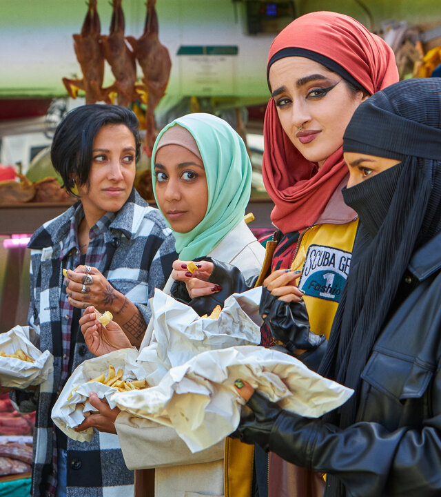 "We are Lady Parts - Bad Girls des Islam": Saira (Sarah Kameela Impey), Amina (Anjana Vasan), Ayesha (Juliette Motamed) und Momtaz (Lucie Shorthouse) stehen an einem Marktstand und essen gemeinsam Pommes. Im Hintergrund sind Fleischwaren in der Auslage zu sehen.