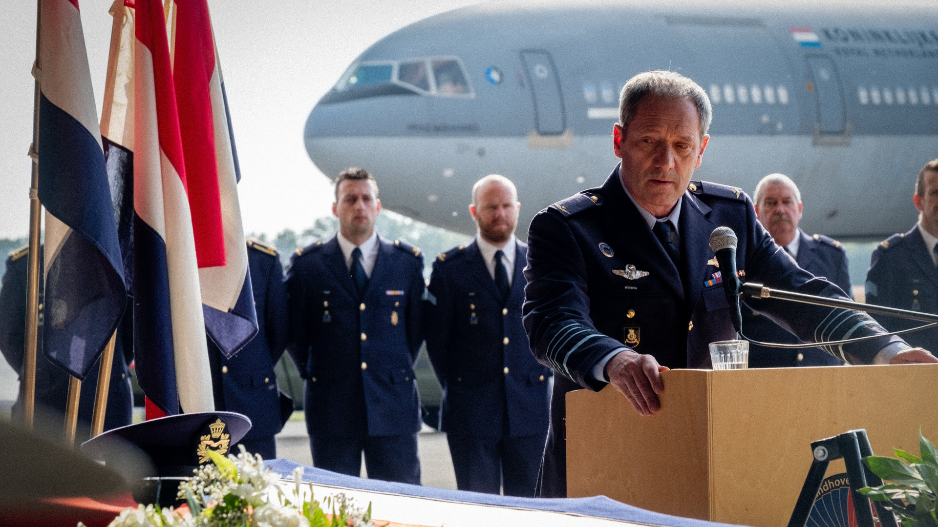"High Flyers - Panda-Diplomatie": Roderick Walema (Gijs Scholten van Aschat) steht mit traurigem Gesichtsausdruck hinter einem Pult mit Mikrofon. Vor ihm steht ein Sarg mit Blumen und niederländischen Flaggen.