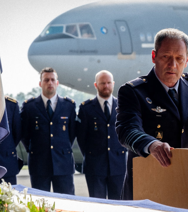 "High Flyers - Panda-Diplomatie": Roderick Walema (Gijs Scholten van Aschat) steht mit traurigem Gesichtsausdruck hinter einem Pult mit Mikrofon. Vor ihm steht ein Sarg mit Blumen und niederländischen Flaggen.