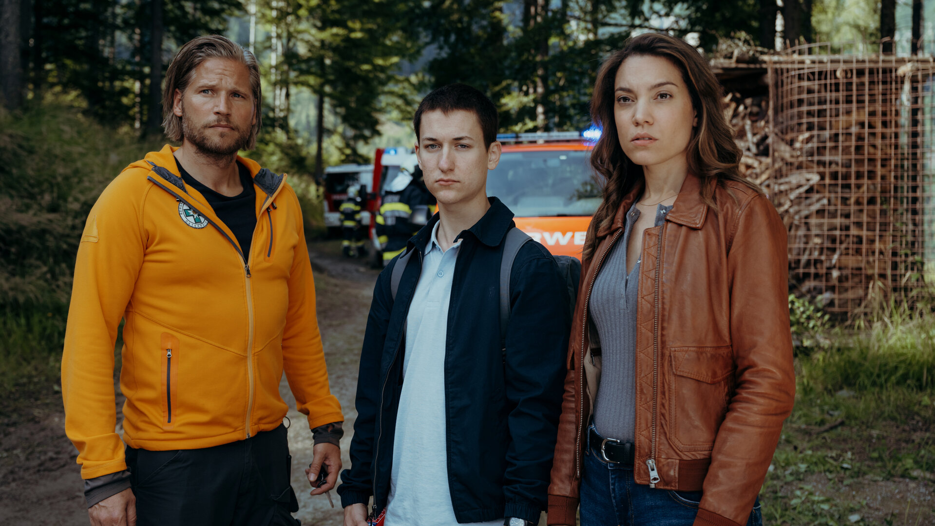 "Die Bergretter: Höhenfeuer": Auf einem Waldweg: Bastian Kessler (Juri Winkler) steht zwischen Alexandra (Viktoria Ngotsé) und dem Bergretter Markus (Sebastian Ströbel). Sie blicken erwartungsvoll in die Ferne. Im Hintergrund stehen Feuerwehrautos.