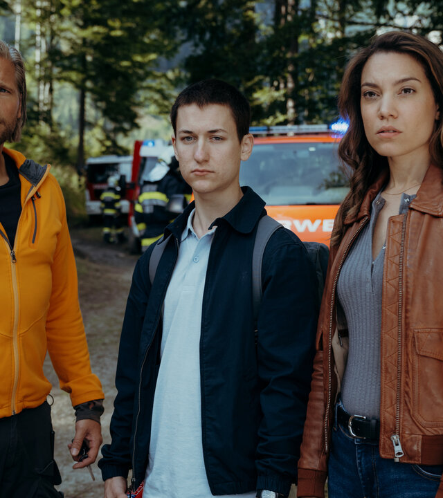 "Die Bergretter: Höhenfeuer": Auf einem Waldweg: Bastian Kessler (Juri Winkler) steht zwischen Alexandra (Viktoria Ngotsé) und dem Bergretter Markus (Sebastian Ströbel). Sie blicken erwartungsvoll in die Ferne. Im Hintergrund stehen Feuerwehrautos.