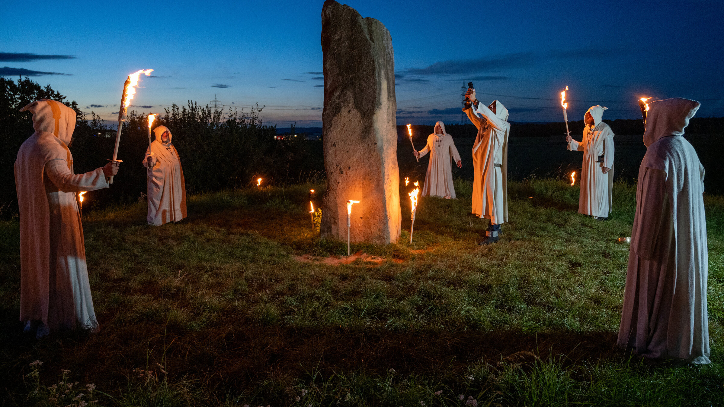 "SOKO Stuttgart - Das Keltengrab": Eine Gruppe Männer in weißen Kutten (Komparsen) steht mit über den Kopf gezogenen Kapuzen und brennenden Fackeln um einen großen Monolithen auf einem Hügel. Neben dem Stein steht der Anführer der Gruppe (Moritz Otto) und streckt einen Kelch zum Himmel.