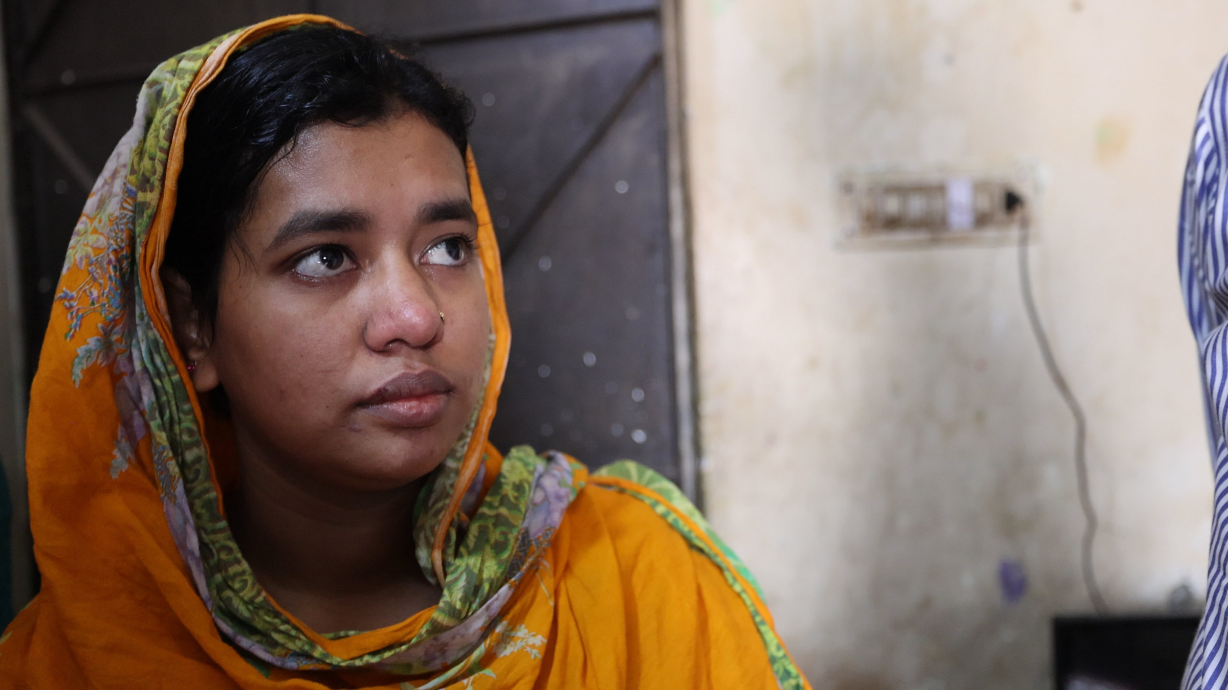 "37°: Fast Fashion - Billige Kleidung für Deutschland": Shimu, eine junge Frau sitzt links im Bild und schaut rechts an der Kamera vorbei. Sie trägt ein orangefarbenes Tuch, das Kopf und ihre Schultern bedeckt. Im Hintergrund sieht man den Teil einer grauen Tür aus Holz und eine helle Lehmwand.