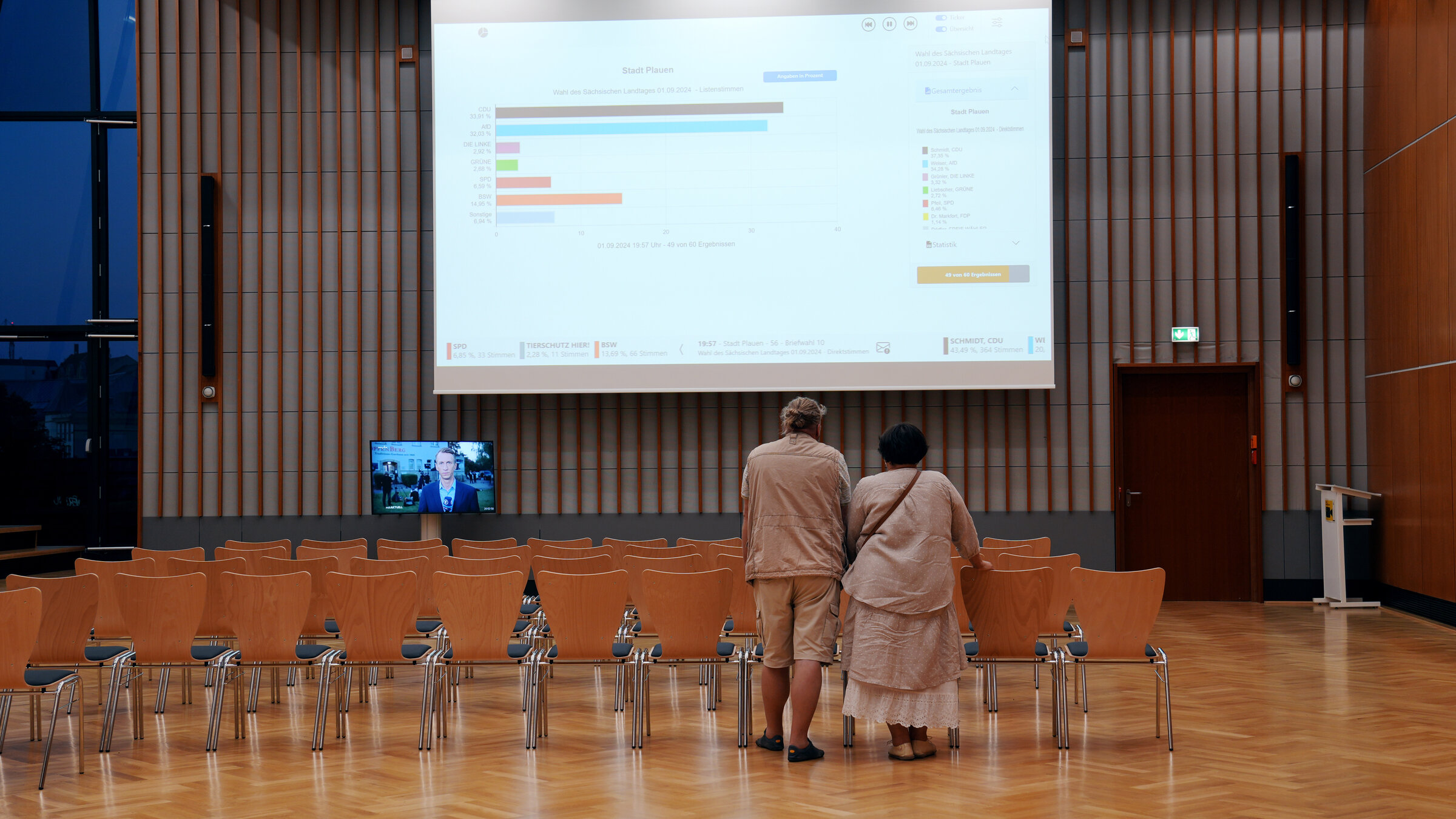 "Drei Frauen gegen Rechtsextremismus": Doritta steht mit ihrem Mann in einem großen Sitzungssaal und schaut auf einen großen Bildschirm.