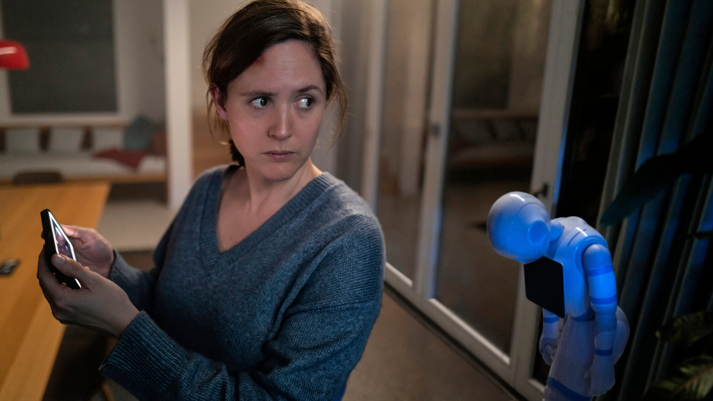 "Unsichtbarer Angreifer": Es ist dunkel im Haus und Emma (Emily Cox) ist offenbar allein. Auch Hausroboter Samy steht ausgeschaltet in der Ecke. Der Bildschirm auf Emmas Handy leuchtet, sie schaut besorgt um sich.