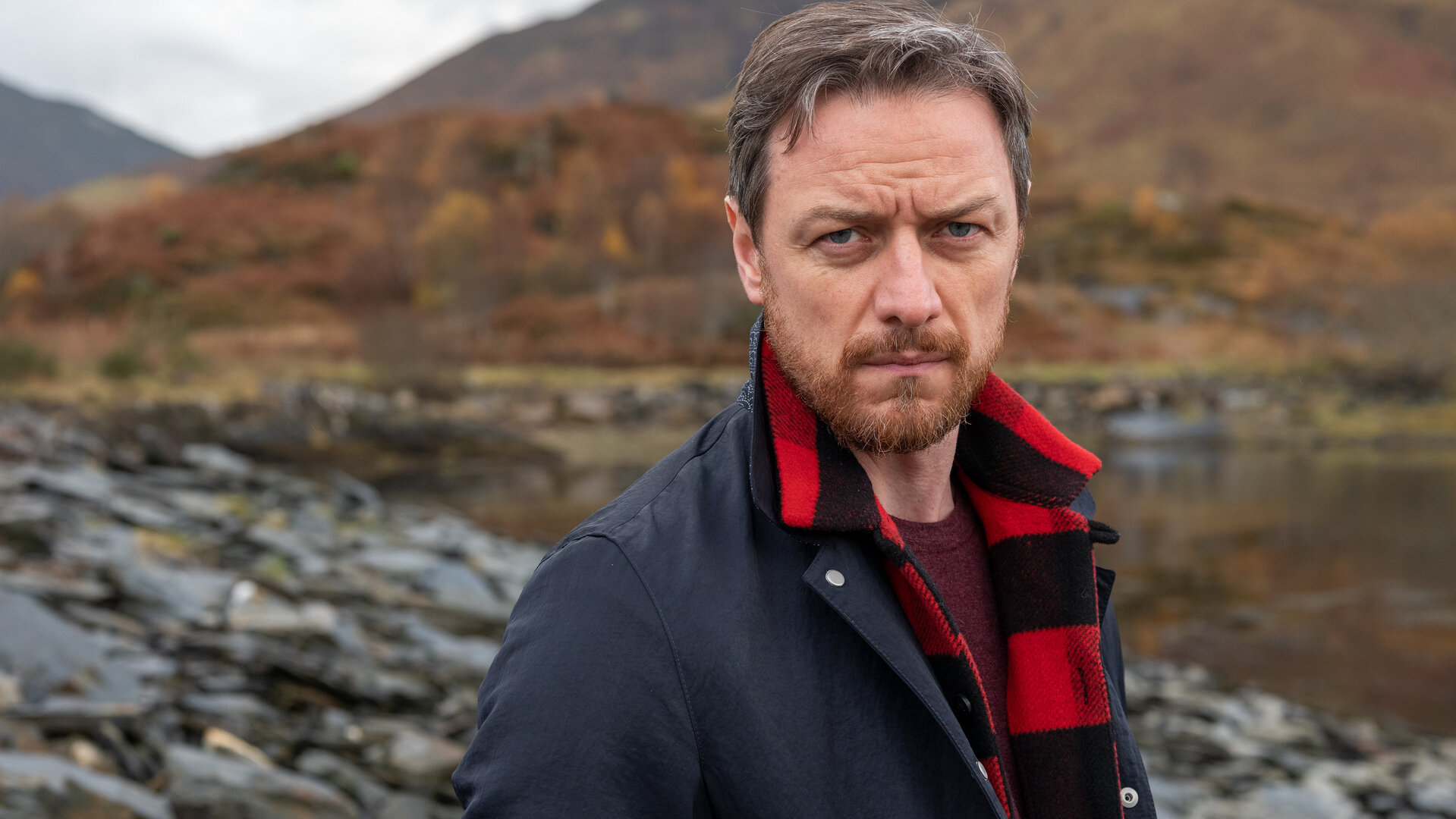 "Vermisst in den Highlands": Edmond Murray (James McAvoy) steht vor einem herbstlichen schottischen Gewässer, einem Loch, und blickt entschlossen nach vorne.