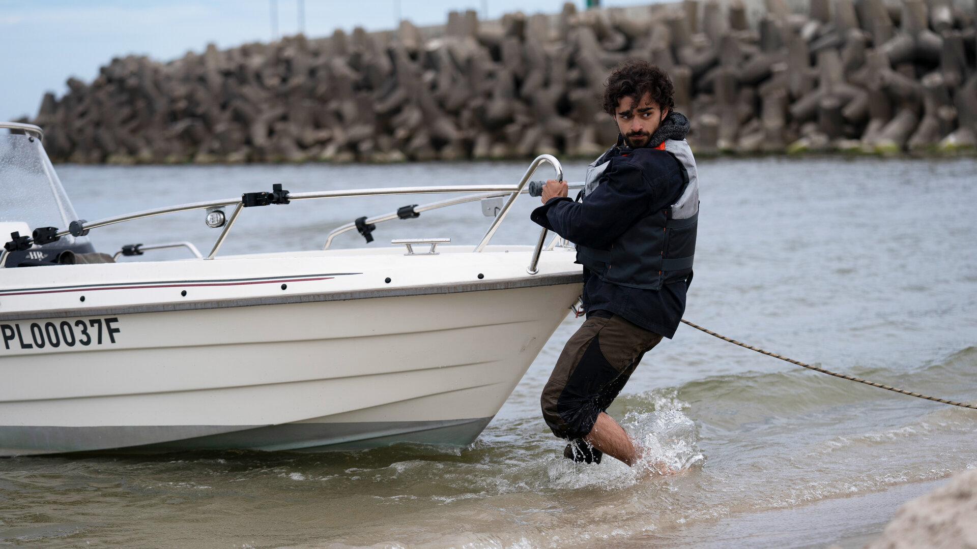 "Sløborn III – Mach dich nützlich": Lobo (Zoran Pingel) steht bis zu den Waden im seichten Wasser und schiebt ein Motorboot vom Strand ins Meer.