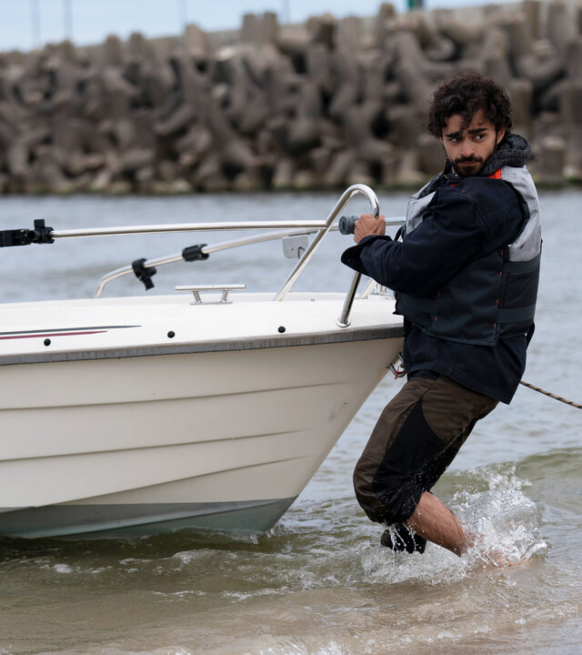 "Sløborn III – Mach dich nützlich": Lobo (Zoran Pingel) steht bis zu den Waden im seichten Wasser und schiebt ein Motorboot vom Strand ins Meer.