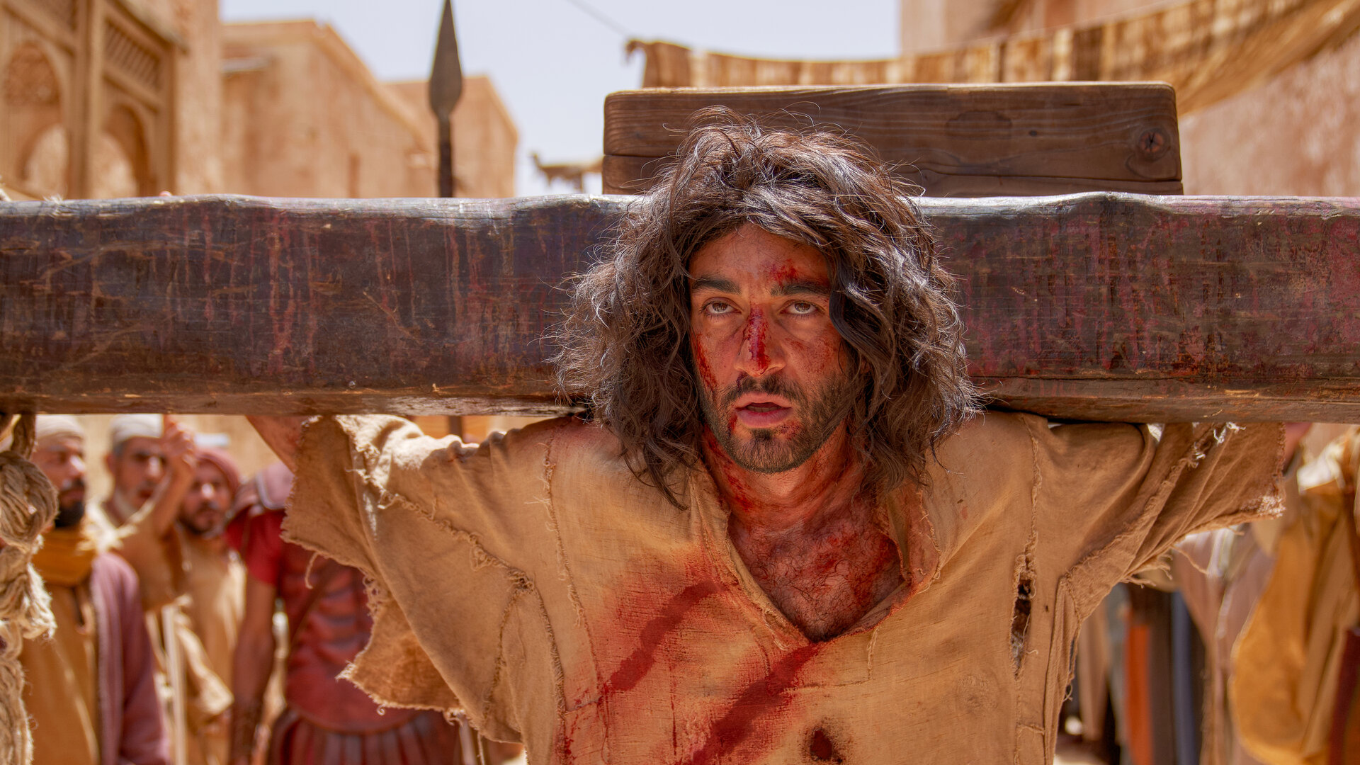 "INRI - Warum musste Jesus sterben?": Das Gesicht von Jesus (Eidin Jalali)  ist von Blutspuren gezeichnet. Dahinter liegt der Querbalken des Kreuzes. Im Hintergrund stehen Menschen.