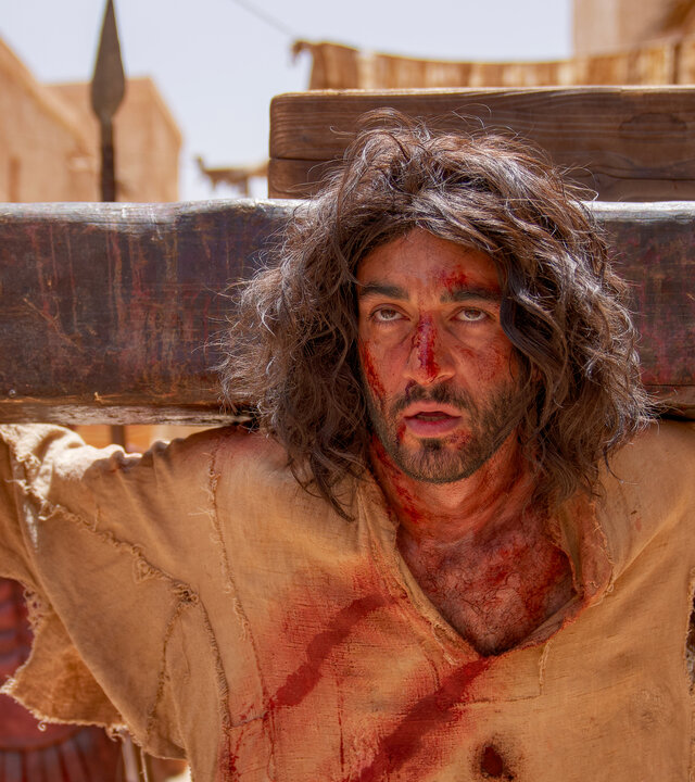 "INRI - Warum musste Jesus sterben?": Das Gesicht von Jesus (Eidin Jalali)  ist von Blutspuren gezeichnet. Dahinter liegt der Querbalken des Kreuzes. Im Hintergrund stehen Menschen.