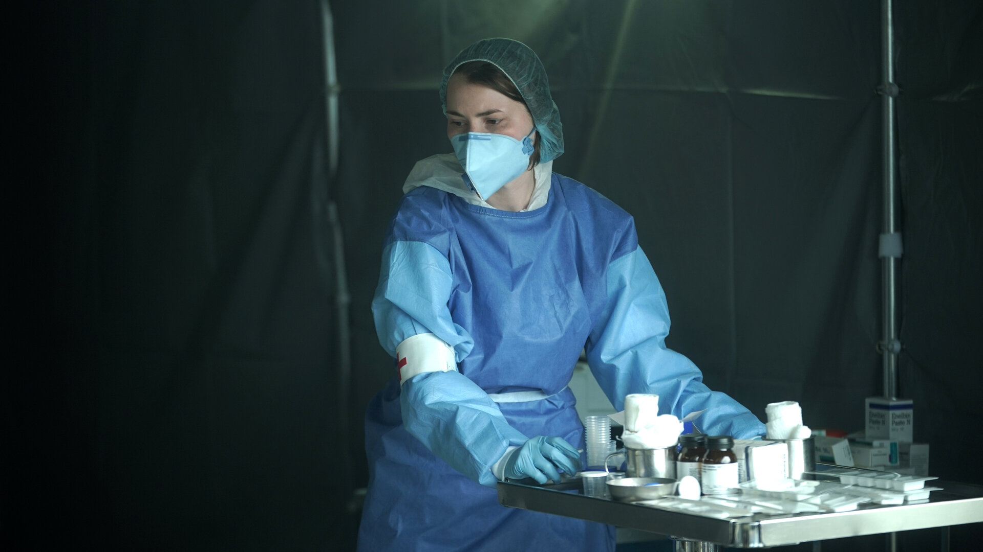 "Sløborn III – Klabaster sterben einsam": Yvonne (Linda Stockfleth) steht in voller Schutzkleidung im Krankenhauszelt vor einem kleinen Metalltisch mit Medikamenten und Verbandszeug.