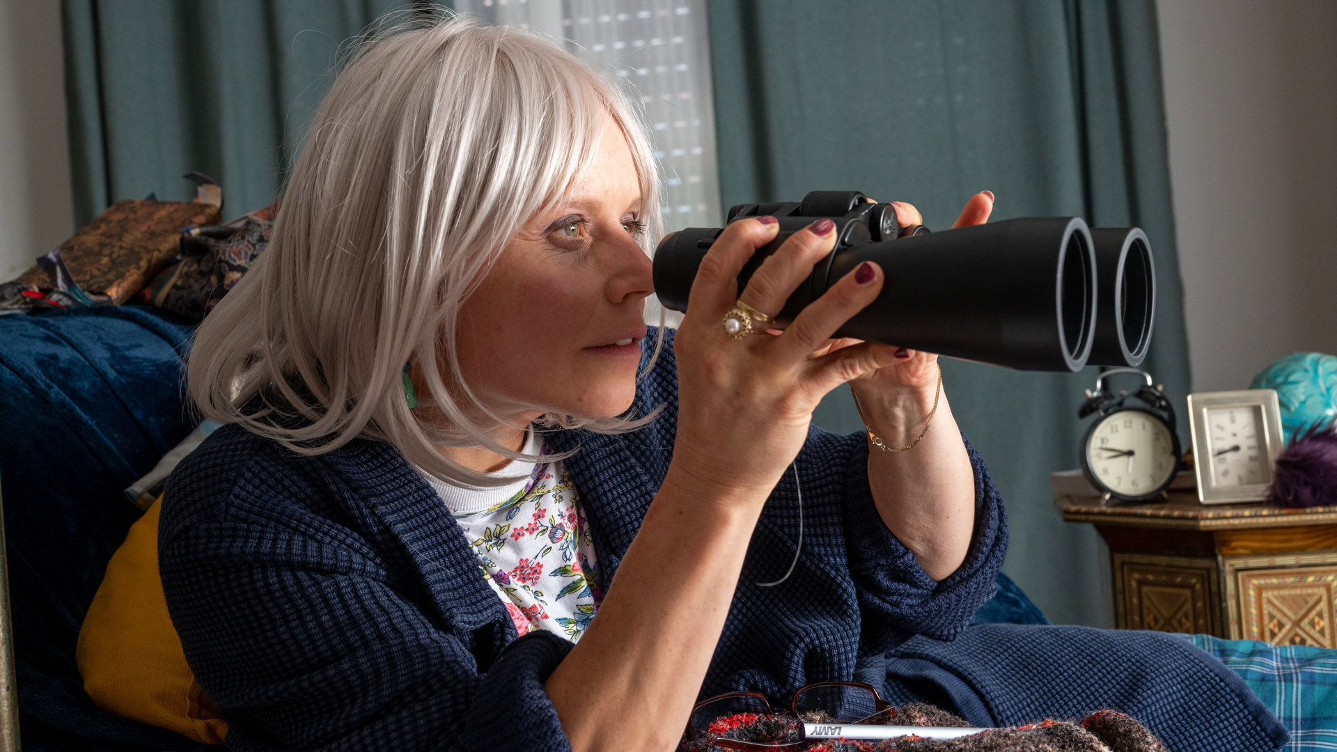"SOKO Stuttgart - Die Frau am Fenster": Alice Seller (Annette Strasser) sitzt in ihrer Wohnung auf einer Couch. Sie hält ein Fernglas in beiden Händen und schaut gespannt durch dieses hindurch. Hinter ihr sieht man auf einem Beistelltisch zwei Wecker nebeneinander stehen. Die Jalousien und die Vorhänge sind zugezogen.