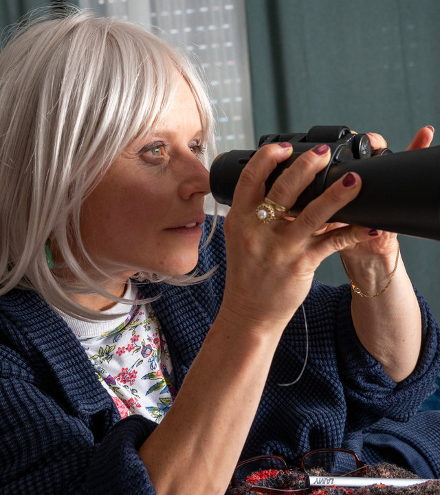 "SOKO Stuttgart - Die Frau am Fenster": Alice Seller (Annette Strasser) sitzt in ihrer Wohnung auf einer Couch. Sie hält ein Fernglas in beiden Händen und schaut gespannt durch dieses hindurch. Hinter ihr sieht man auf einem Beistelltisch zwei Wecker nebeneinander stehen. Die Jalousien und die Vorhänge sind zugezogen.