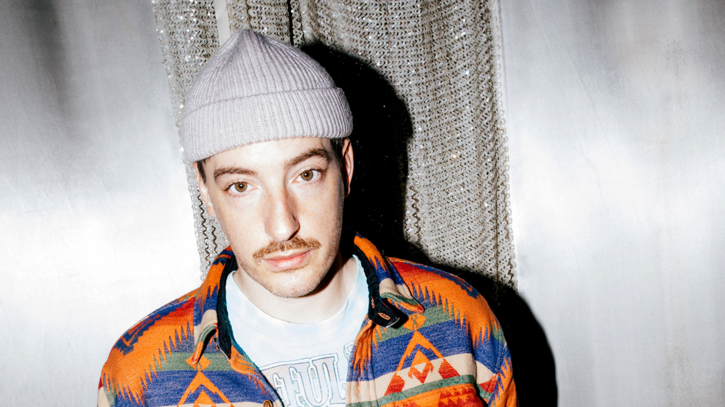 "Casper: Alles war schön und nichts tat weh": Ein junger Mann mit orange-blau gemusterter Jacke und hellgrauer Mütze steht vor einer grauen Wand und schaut direkt in die Kamera.