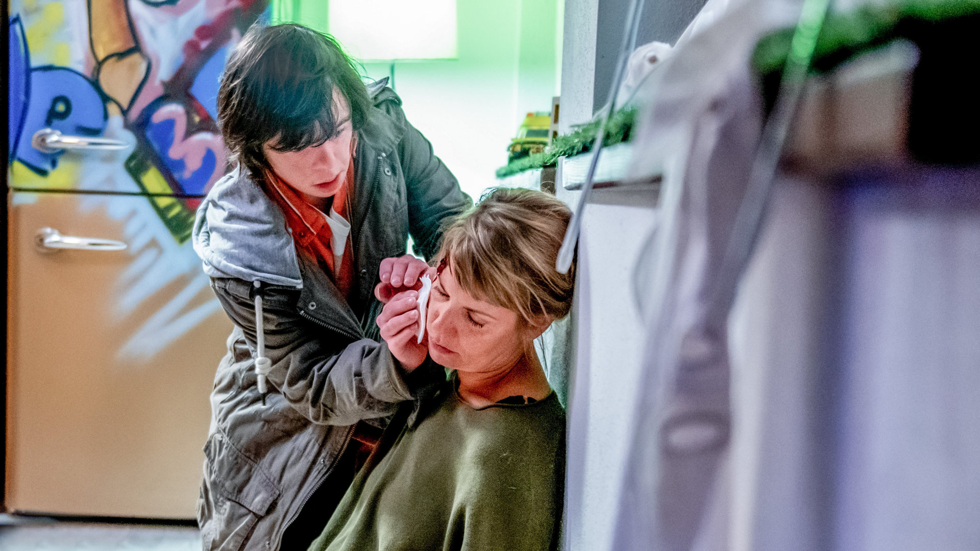 "SOKO Kitzbühel - Allein": In der Küche sitzt Rita (Jaschka Lämmert) auf dem Boden an einem Regal angelehnt. Sie blutet an der Schläfe. Ihr Sohn Marius (Brent Larsen) hält entsetzt ein Taschentuch an die Wunde, um die Blutung zu stoppen.