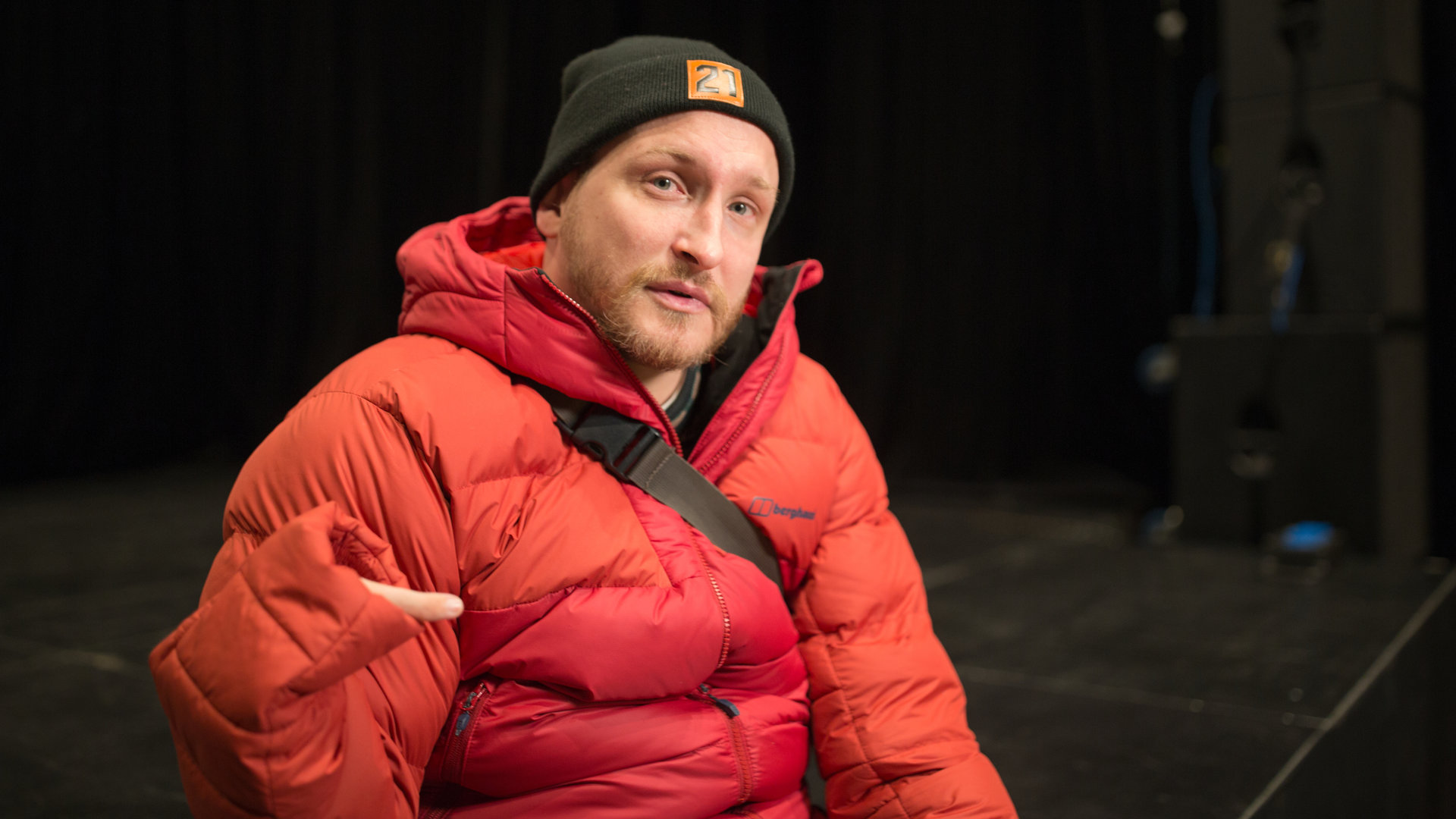 "einfach Mensch: Graf Fidi: Ich mach das mit links": Graf Fidi mit roter Daunenjacke und einer dunklen Mütze auf einer Bühne.