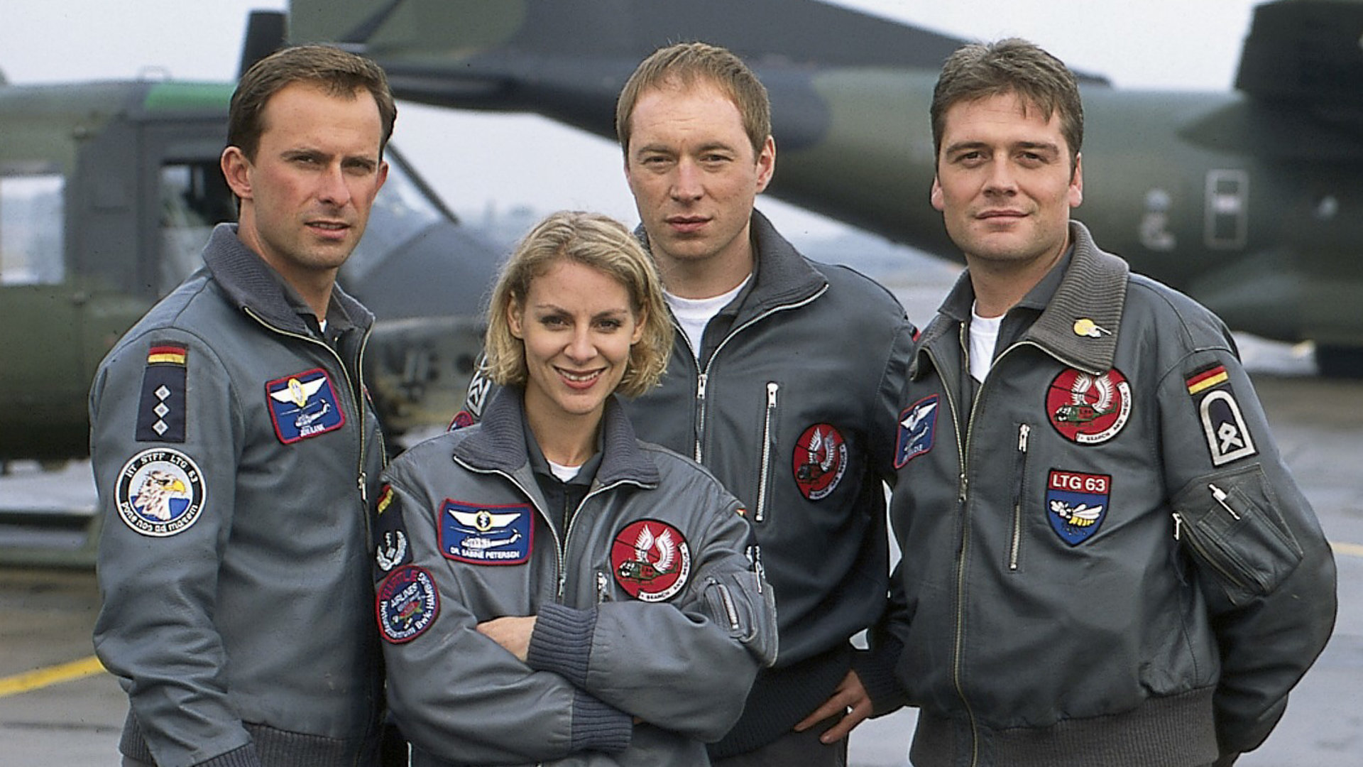 "Die Rettungsflieger - Rivalen im Cockpit": Teamaufnahme von Jens Blank (Nicolas König), Sabine Petersen (Marlene Marlow), Paul Reinders (Rainer Sellien) und Jan Wollcke (Oliver Hörner) auf dem Flugplatz. Im Hintergrund ein Transportflugzeug und ein Hubschrauber.