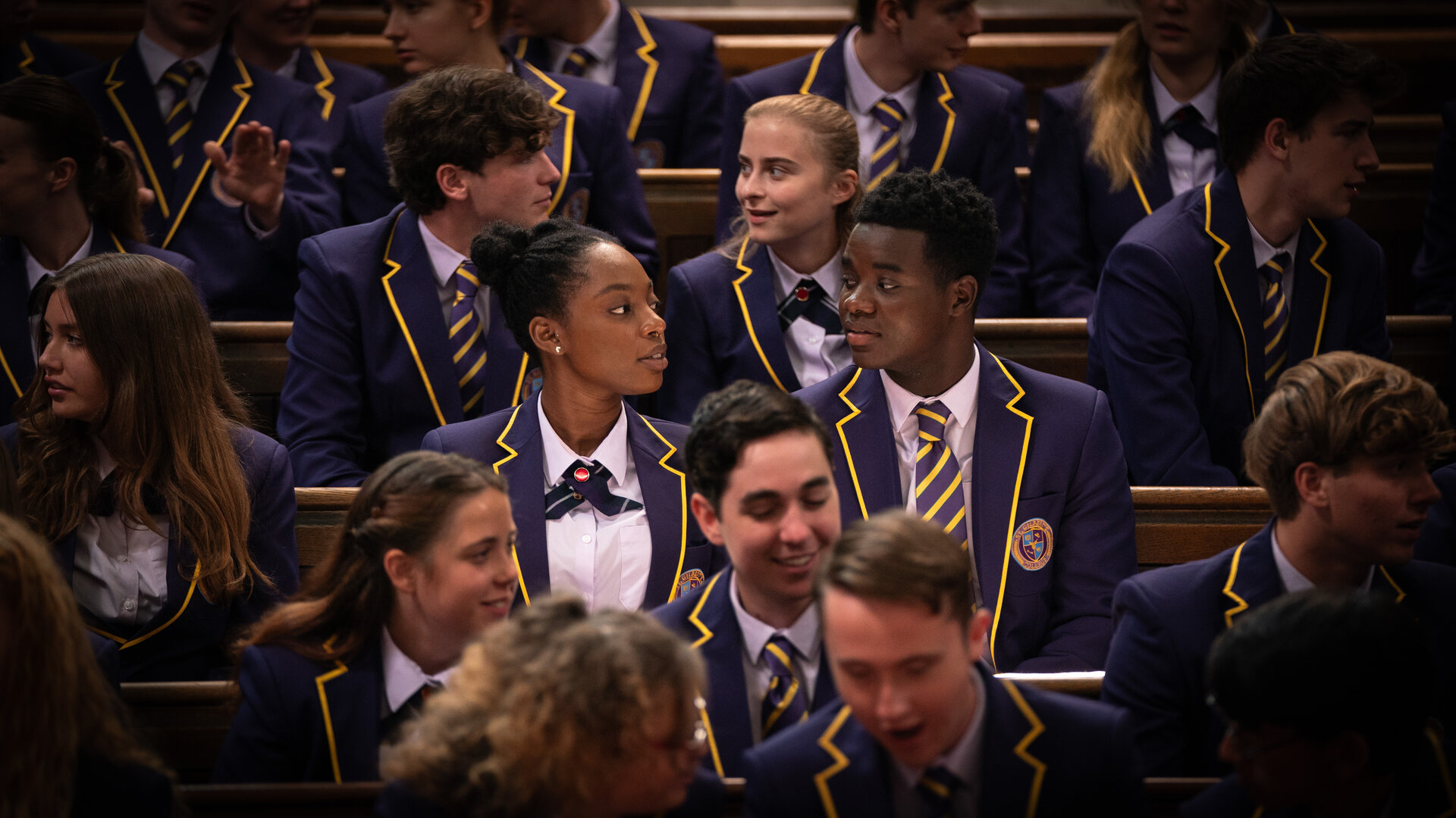 "Boarders - Welcome to St. Gilbert's - Police & Paintball": Leah (Jodie Campbell) und Jaheim (Josh Tedeku) sitzen nebeneinander auf einer vollen Tribüne. Sie tragen ihre Schuluniformen.