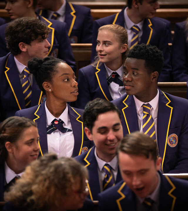 "Boarders - Welcome to St. Gilbert's - Police & Paintball": Leah (Jodie Campbell) und Jaheim (Josh Tedeku) sitzen nebeneinander auf einer vollen Tribüne. Sie tragen ihre Schuluniformen.