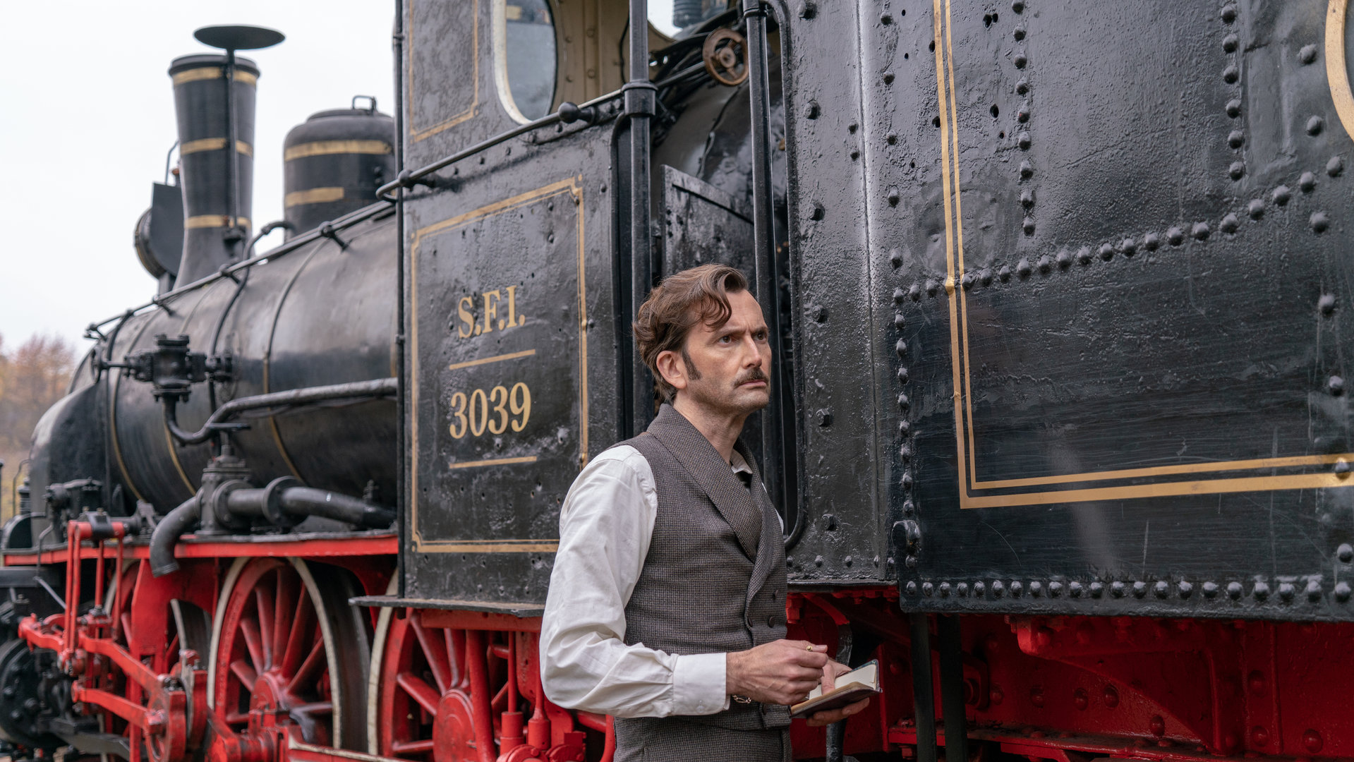 "In 80 Tagen um die Welt (2)": Phileas Fogg (David Tennant) steht seitlich vor einer Dampflokomotive und hält ein Notizbuch in seiner Hand.