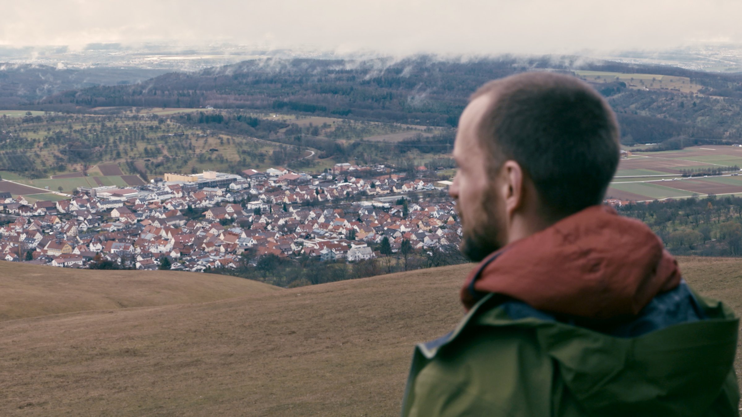 "Die Karte der Schönheit": Vor dem Panorama einer hügeligen Landschaft, in der zwischen Grünflächen und Waldstücken auch Siedlungen zu erkennen sind, steht ein schlanker Mann mit Vollbart und grünem Anorak und schaut in die Ferne. Man sieht ihn von schräg links hinten.