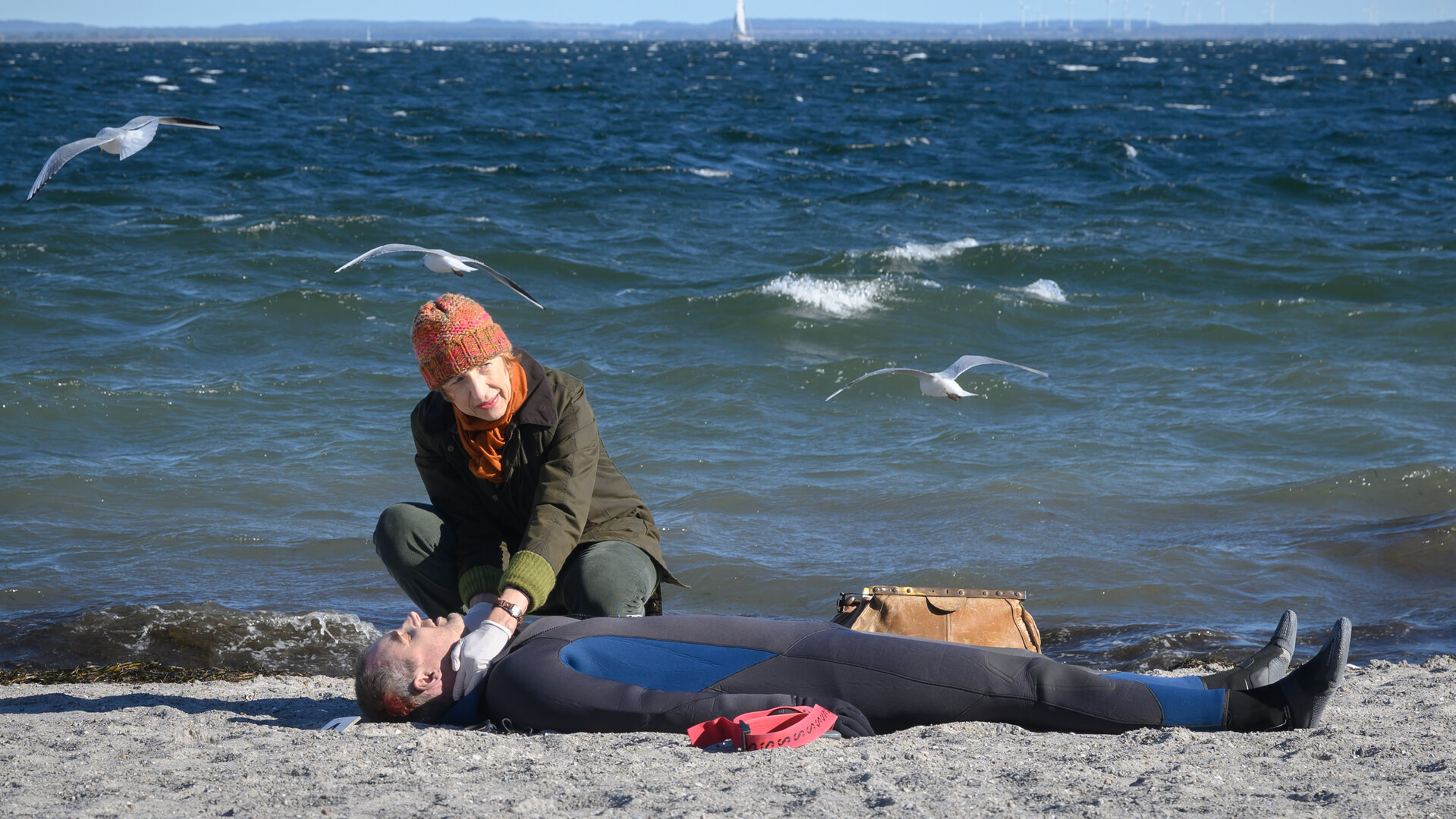 "SOKO Wismar – Die andere Hälfte": Rechtsmedizinerin Dr. Helene Sturbeck (Katharina Blaschke) untersucht am Strand die Leiche von Apnoetaucher Sören Hunke (Sönke Schnitzer), der einen Neoprenanzug trägt und auf dem Rücken im Sand liegt. Im Hintergrund liegt die Ostsee unter einem blauen Himmel, zwei Möwen fliegen über dem Wasser.