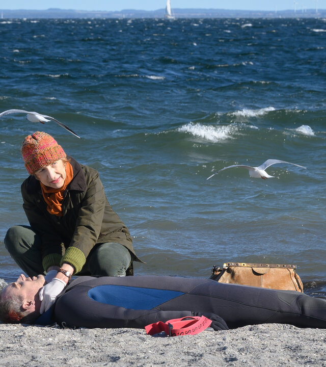 "SOKO Wismar – Die andere Hälfte": Rechtsmedizinerin Dr. Helene Sturbeck (Katharina Blaschke) untersucht am Strand die Leiche von Apnoetaucher Sören Hunke (Sönke Schnitzer), der einen Neoprenanzug trägt und auf dem Rücken im Sand liegt. Im Hintergrund liegt die Ostsee unter einem blauen Himmel, zwei Möwen fliegen über dem Wasser.