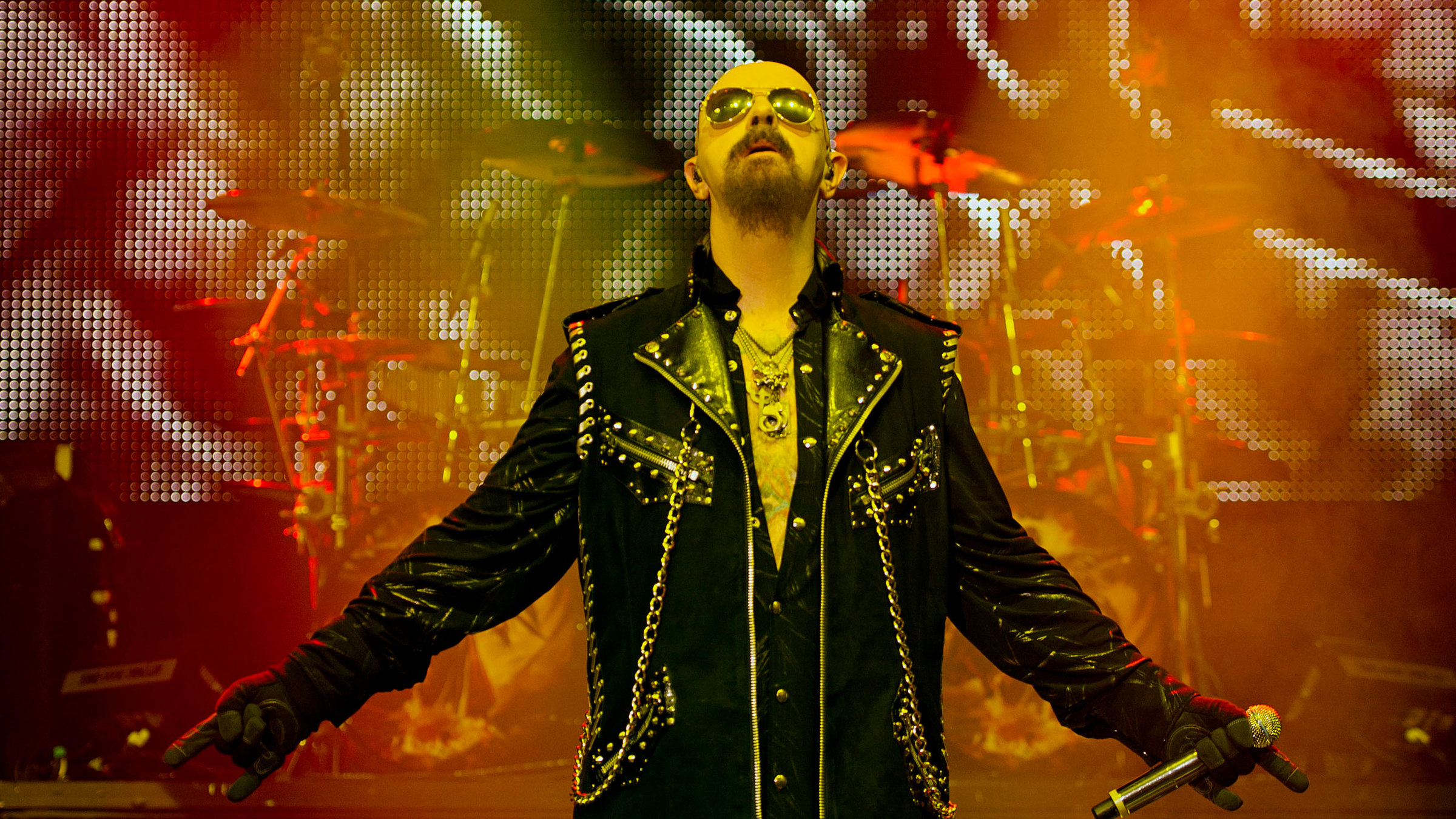 "Pop Around the Clock – Summer Edition: Judas Priest: Battle Cry - Live at Wacken": Ein Mann mit schwarzer Sonnenbrille und Glatze steht auf einer Bühne mit bunter LED-Lichterwand im Hintergrund. Er trägt eine schwarze Lederhose und eine mit silbernen Nieten besetzte schwarze Lederjacke. Er hat die Arme unten seitlich ausgebreitet und schaut nach oben.