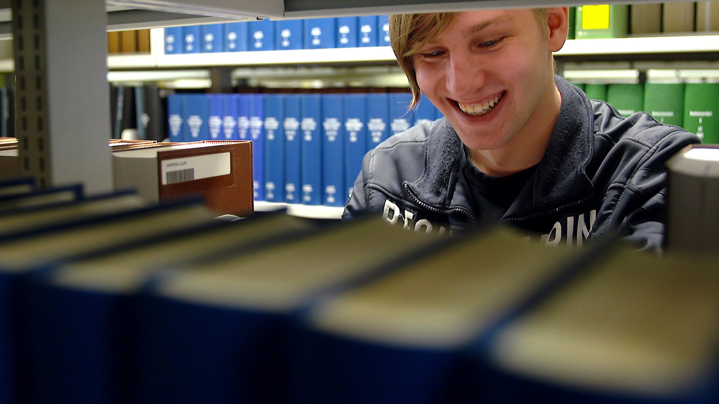 "37° Die Beginner (1) - Vom aufregenden Start ins Leben": Dennis steht lachend zwischen zwei Bücherregalen in der Uni-Bibliothek.