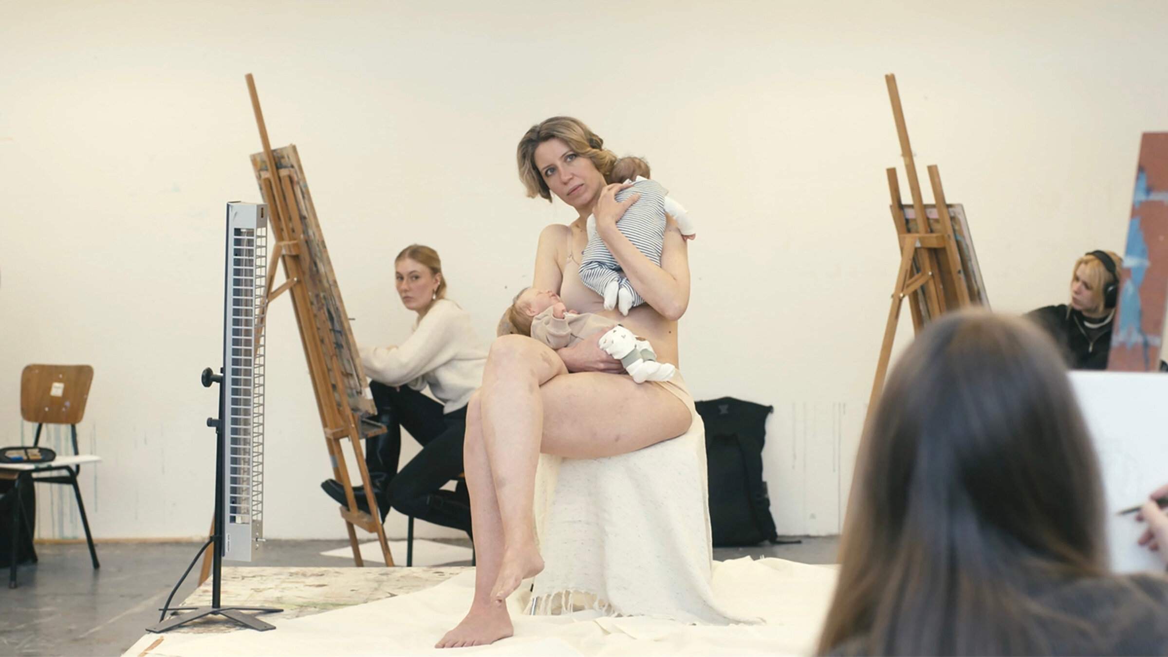 "Reproduktion": Sitzend posierende Frau mit Babypuppen auf dem Schoß und im Arm, umringt von Malenden an Staffeleien.