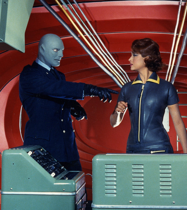 "Fantomas": Fantomas (Jean Marais), Lady Beltham (Marie-Hélène Arnaud) und sein Leibwächter (Henri Attal) stehen in einem roten Raum vor einigen technischen Geräten. Während Fantomas energisch auf ein Gerät zeigt, schaut Lady Beltham ihn direkt an und der Leibwächter schaut konzentriert nach unten.