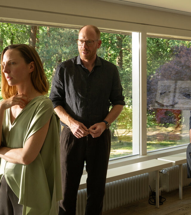 "Wendland - Stiller und der Teufelssauger": Jakob Stiller (Ulrich Noethen), Oda Nolde (Katharina Schüttler) und Jan Nolde (Johann von Bülow) stehen zusammen in einem Wohnzimmer. Oda Nolde steht mit dem Rücken zu Jakob Stiller und Jan Nolde. Hinter ihnen ist der Garten durch eine Fensterfront zu sehen.