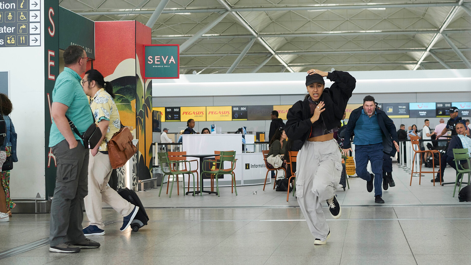 "The Outlaws - Theorie und Praxis": Polizist Burgess (Ricky Grover) rennt auf einem Flughafen hinter Rani (Rhianne Barreto) her. Alle Passanten (Komparsen) drehen sich nach ihnen um.