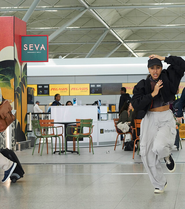 "The Outlaws - Theorie und Praxis": Polizist Burgess (Ricky Grover) rennt auf einem Flughafen hinter Rani (Rhianne Barreto) her. Alle Passanten (Komparsen) drehen sich nach ihnen um.