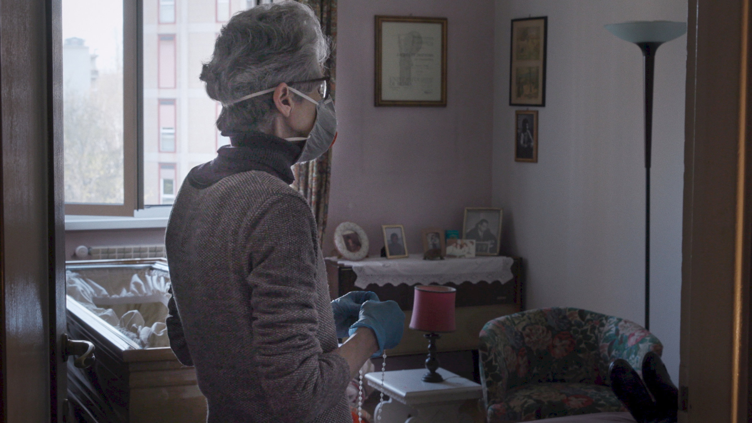 "Die Welt jenseits der Stille": Eine schlanke Frau mit kurzen grauen Haaren und grauem Pullover, die eine Mund-Nasen-Bedeckung trägt, steht, von der Kamera abgewendet, in einem Zimmer und betet den Rosenkranz. Links neben ihr steht ein offener Sarg.