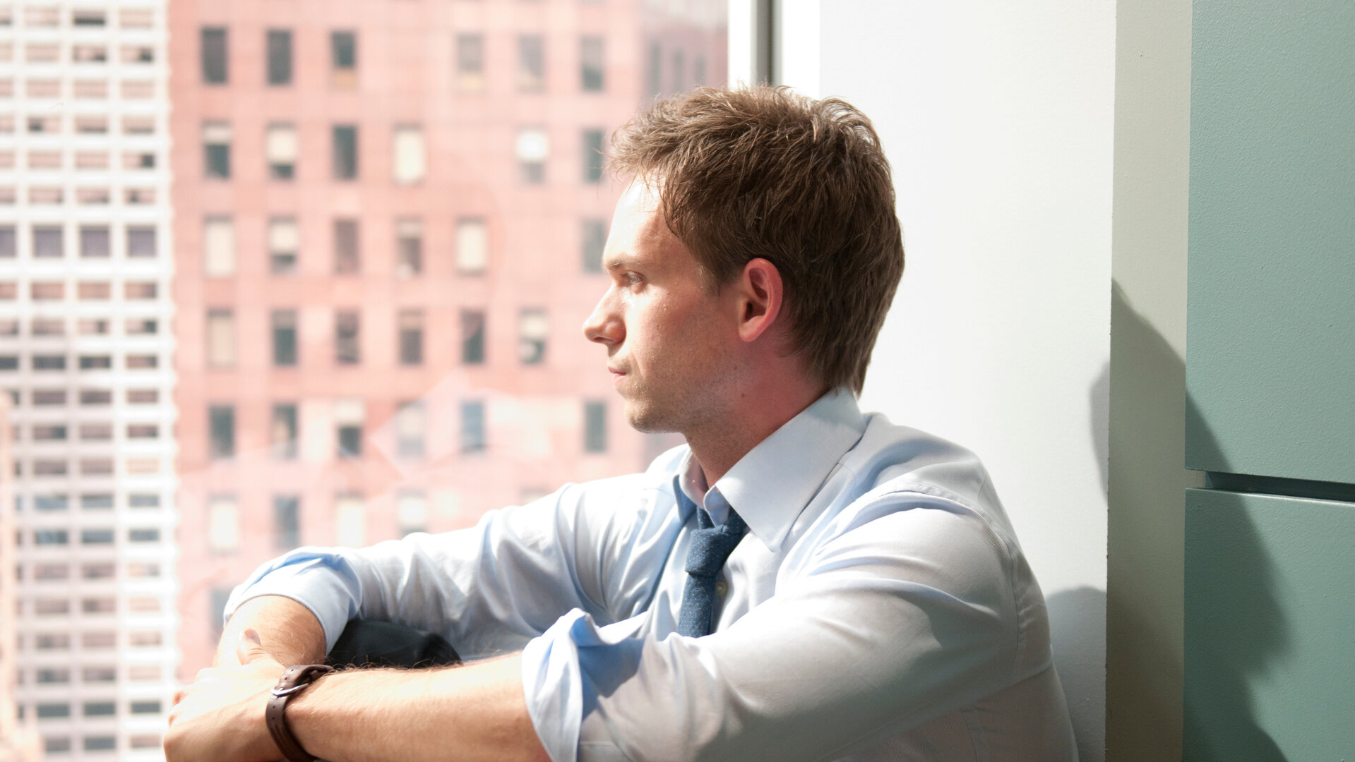 "Suits - Überholmanöver": Mike (Patrick J. Adams) sitzt in einem Hemd mit hochgekrempelten Ärmeln vor einem großen Fenster. Er blickt nachdenklich in die Ferne, das Licht fällt hell auf sein Gesicht, im Hintergrund ist die Fassade eines Hochhauses zu sehen.